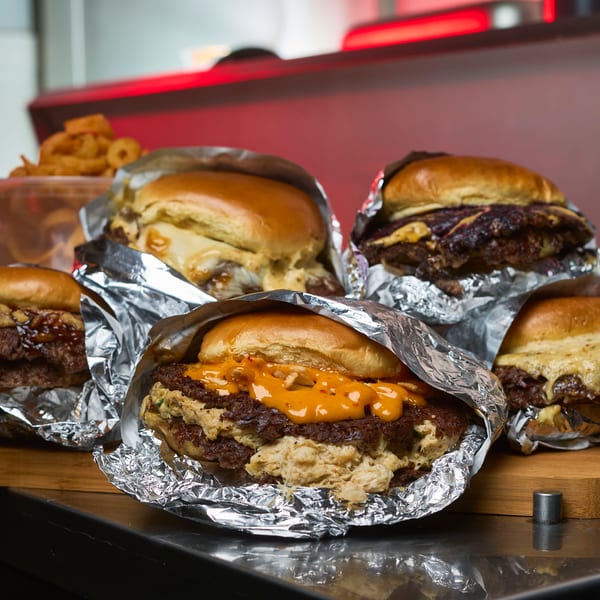 Multiple smash burgers wrapped in foil with crispy edges and sauces on a wooden board.