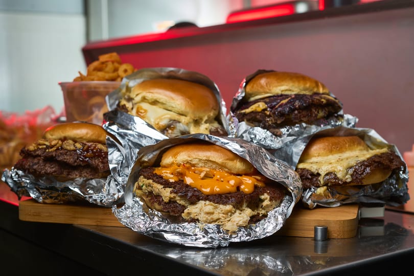 Multiple smash burgers wrapped in foil with crispy edges and sauces on a wooden board.