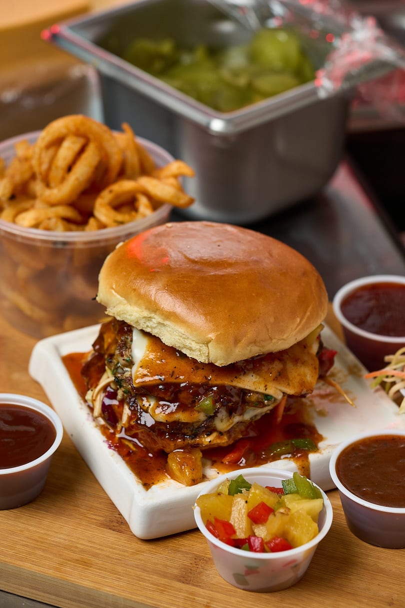 Loaded jerk burger with pineapple salsa, curly fries, and BBQ sauces on a wooden board.