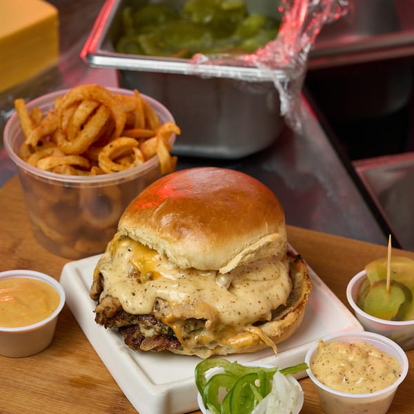Cheesy smash burger with jalapenos, pickles, curly fries, and spicy sauces on a wooden board.