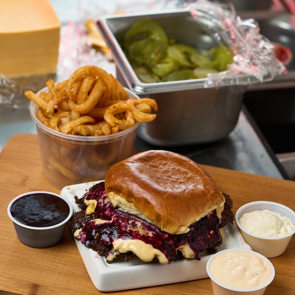 Blueberry smash burger with berry compote, curly fries, and dipping sauces on a wooden board.