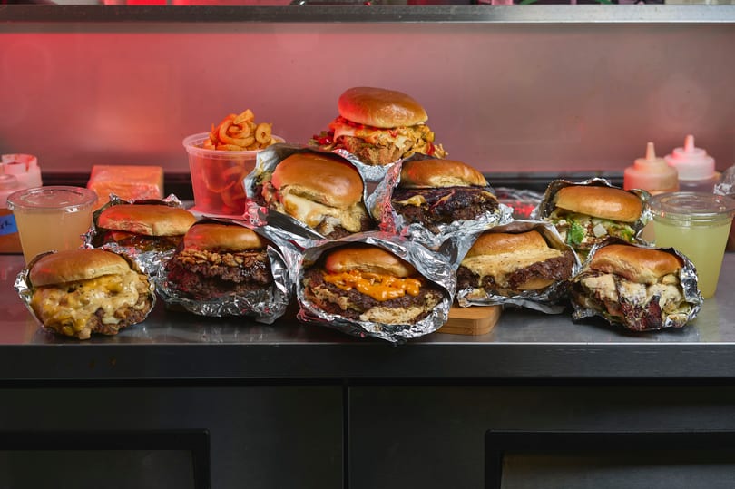 Full spread of smash burgers wrapped in foil with curly fries and lemonade on a counter.