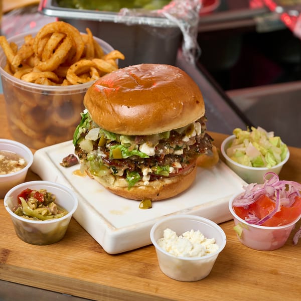 Greek-style smash burger with feta, lettuce, pickled onions, and curly fries on a cutting board.