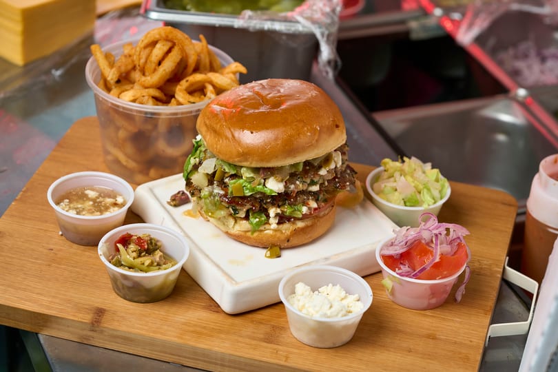 Greek-style smash burger with feta, lettuce, pickled onions, and curly fries on a cutting board.